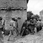 A platoon of black troops surround a house as they prepare to eliminate a German sniper holding up an advance, on Omaha Beach, near Vierville Sur-Mer, France. (June 10, 1944). Source: U.S. National Archives, # 111-SC-190120.