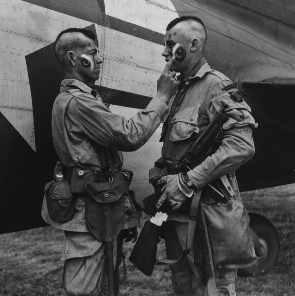 Private Clarence C. Ware applies last second make-up to Private  Charles R. Plaudo (members of the 101st Airborne Division) in England before D-Day. June 1944. Source: U.S. National Archives, # 111-SC-193551.