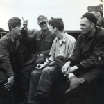 Aboard a Coast Guard-manned LCI bound from the French coast to England, Nazi prisoners give a U.S. Coast Guardsman the lowdown on how they happened to be captured. German prisoner with his finger pointing upward is translating his colleague's remarks about Allied air power and the U.S. Coast Guardsman, at his right, is Robert E. O'Connell. Source: U.S. National Archives, # 26-G-2448.