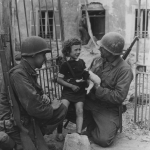 Left to right: Private Roland Bonnell and Sergeant James Devine, both members of an engineer unit take time out to comfort a French girl as U.S. troops force ahead in France. Colleville, France. (June 13, 1944). Source: U.S. National Archives, # 111-SC-190253.