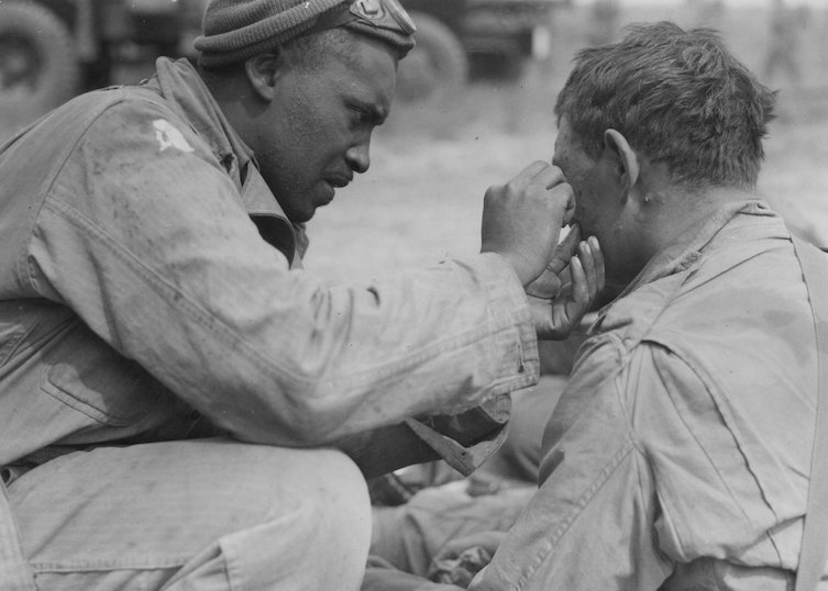 An American soldier lends a hand to another American picking a small bit of metal from his face. Orchard Beach, France. (June 8, 1944). Source: U.S. National Archives, # 111-SC-190258.