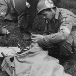 American Medical Officer, who landed with the 82nd Airborne Division in France, hands a lighted cigarette to a fellow officer who had an adverse landing. St. Mere Eglise, France. (June 12, 1944). Source: U.S. National Archives, # 111-SC-190289.