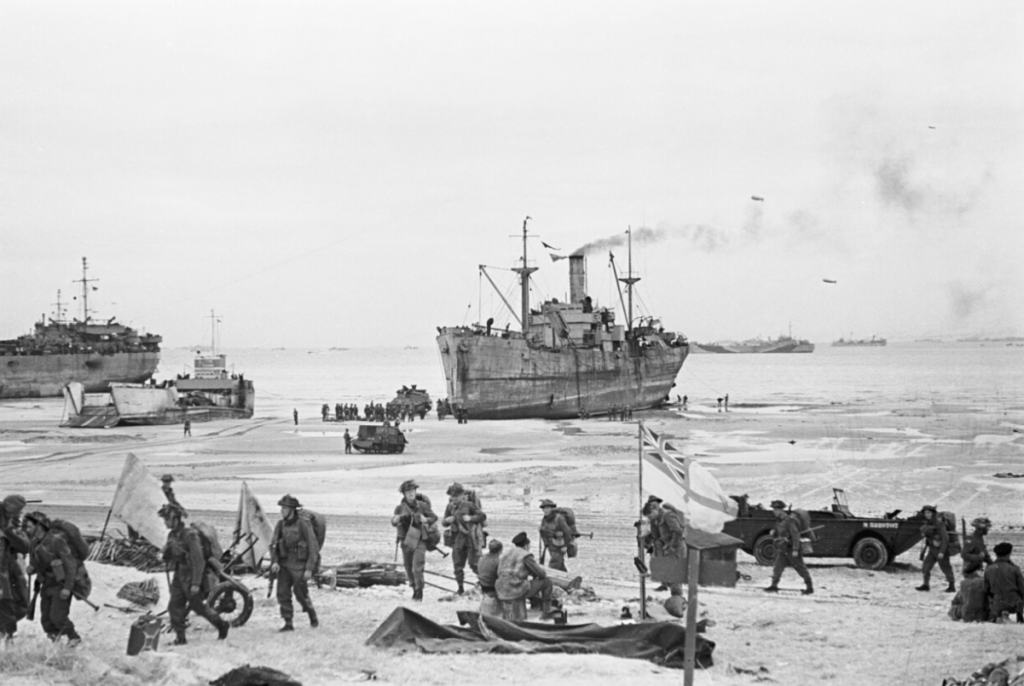 Troops come ashore on one of the Normandy invasion beaches, past the White Ensign of a naval beach party. (June 7, 1944). Source: Imperial War Museums, A 24012.