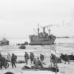 Troops come ashore on one of the Normandy invasion beaches, past the White Ensign of a naval beach party. (June 7, 1944). Source: Imperial War Museums, A 24012.