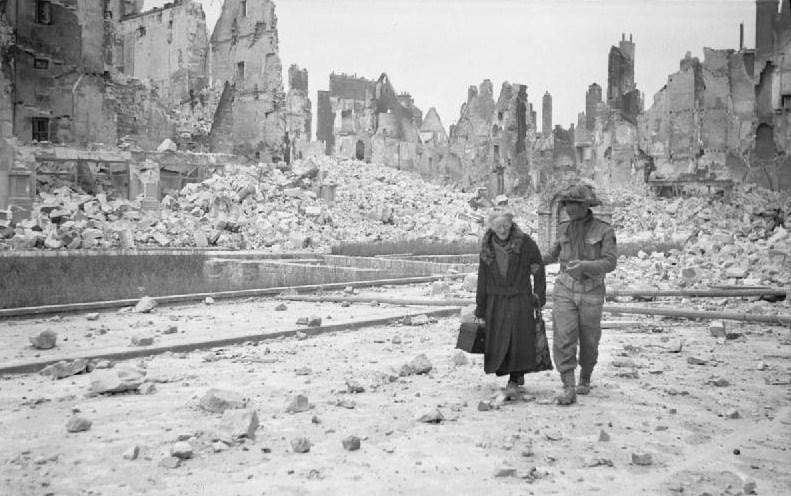A British soldier in Caen gives a helping hand to an old lady amongst the scene of utter devastation. (1944). Source: Imperial War Museums,  B 6794.