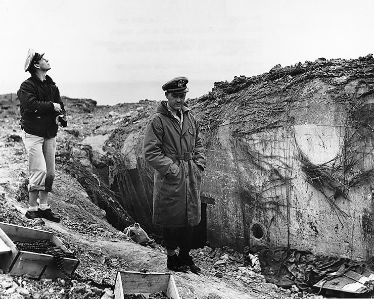 U.S. Navy Lieutenant Commander Knapper and Chief Yeoman Cook of USS Texas examining damaged German pillbox at Pointe du Hoc, Normandy, France. Covered dead U.S. Army Ranger at right. (June 6, 1944). Source: U.S. National Archives, # 80-G-235595.