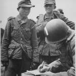 Young Japanese man wearing a Nazi uniform in a round-up of German prisoners on the beaches of France, gives his name and number to an American army captain. Source: U.S. National Archives, 513174.